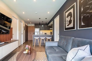 Ein modernes Wohnzimmer mit einem grauen Sofa, einem Couchtisch mit Blumen, einer Küche im Hintergrund und Kunstwerken an den Wänden.