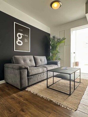 A modern living room with a grey sofa, a coffee table, a large plant, and a framed artwork on the wall.