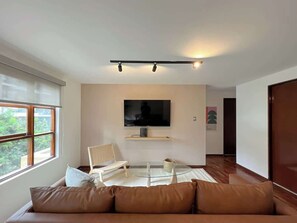Una sala moderna con un sofá, una mesa de centro de vidrio, un televisor montado en la pared y una ventana con vistas a zonas verdes.