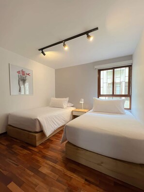 Una habitación con dos camas, piso de madera, una ventana y una lámpara en el techo.