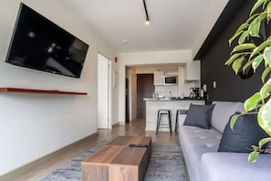 A modern living room with a flat-screen TV, a sofa, a coffee table, and a kitchen area in the background.
