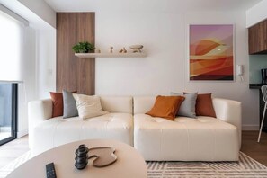 A modern living room with a white leather sofa, a coffee table, and a wall-mounted shelf with decorative items. - Geko | Modern 1BR in San Isidro Central Area (San Isidro)