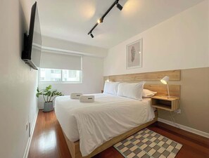 A bedroom with a bed, bedside table, lamp, wall-mounted TV, potted plant, and a window.