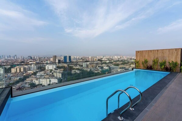 A rooftop pool with a city view, metal pool ladder, and a wooden deck.