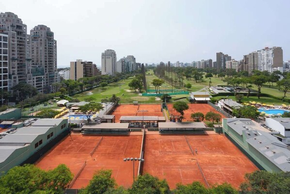 Tenniskenttä, jonka ympärillä on korkeita kerrostaloja, viheralue ja uima-allas.