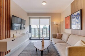 Ein modernes Wohnzimmer mit einer Couch, einem Couchtisch, einem an der Wand befestigten Fernseher und einem großen Fenster mit Blick auf Gebäude.