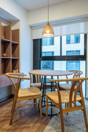 A dining area with a round table and four chairs, a hanging light fixture, and a view of a building through a window with blinds.
