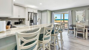 A modern kitchen with a marble countertop, white cabinetry, and stainless steel appliances. There is a dining area with white chairs and a table, and a view of the beach through large windows.