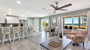 A modern kitchen with white cabinets, a dining area with a table and chairs, a ceiling fan, and a view of the ocean through large windows.