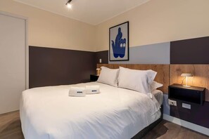 Ein Hotelzimmer mit Bett, weißer Bettwäsche, einem Nachttisch mit Lampe und einem gerahmten Bild an der Wand.