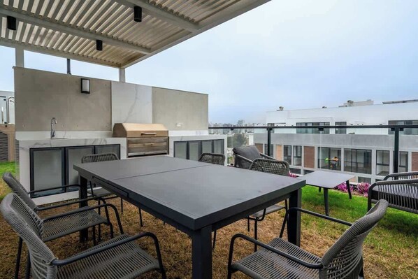 Un patio al aire libre con una mesa, sillas, una parrilla y vistas de edificios al fondo.