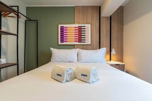 Una habitación de hotel con una cama, dos toallas enrolladas con la marca 'Yekko', una mesita de noche con una lámpara y un cuadro abstracto colorido en la pared.
