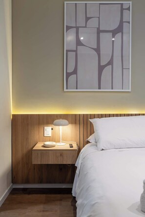 A bedroom with a wooden headboard, a bedside table with a lamp, a framed abstract artwork, and a bed with white bedding.