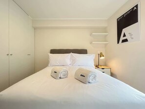 A hotel room with a bed, pillows, a nightstand with a lamp, and a wall-mounted shelf. - Geko | Elegant 1BR w/ Pool near San Isidro (San Isidro)