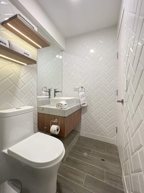 A modern bathroom with a white herringbone tile backsplash, a wooden vanity, a toilet, and a shower area.