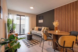 Una sala moderna con un sofá, sillones, una mesa de centro y una mesa de comedor. Hay un balcón con plantas y vistas al exterior.
