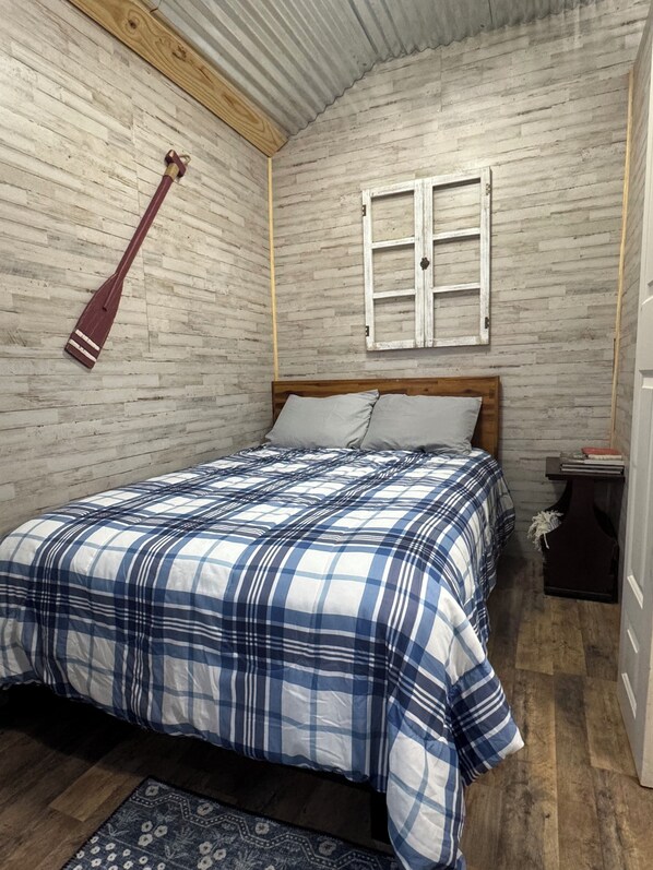 A bedroom with a wooden headboard, a plaid bedspread, a wooden floor, a small bedside table with a book, and a rustic wall with a wooden plank and a metal ceiling.