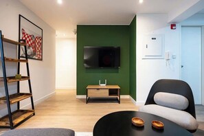 A modern living room with a green accent wall, a mounted TV, a wooden shelf, a black sofa, and a round coffee table. - Geko | Elegant 1BR w/ Free Parking in Barranco (Barranco)