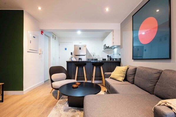A modern living room with a sofa, a round coffee table, and a kitchen area in the background. - Geko | Elegant 1BR w/ Free Parking in Barranco (Barranco)