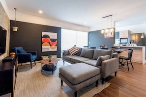 Ein modernes Wohnzimmer mit einer Couch, Hocker und Couchtisch, ein Essbereich mit Tisch und Stühlen, und eine Küche im Hintergrund.