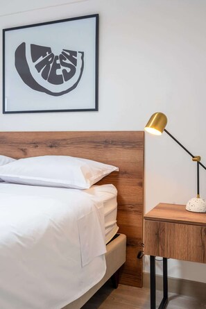 Ein ordentlich bezogenes Bett mit weißen Leinen, ein Holz-Kopfteil, ein gerahmtes abstraktes Kunstwerk darüber und ein Nachttisch mit einer Lampe.