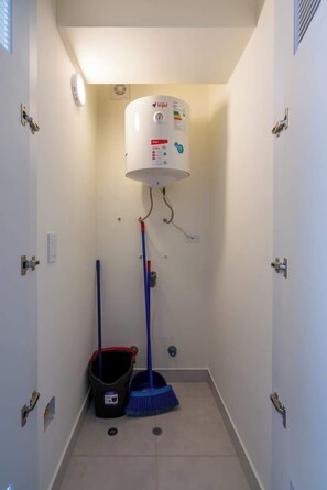 Une salle de bain avec des toilettes blanches, un radiateur fixé au mur, un seau et un balai.