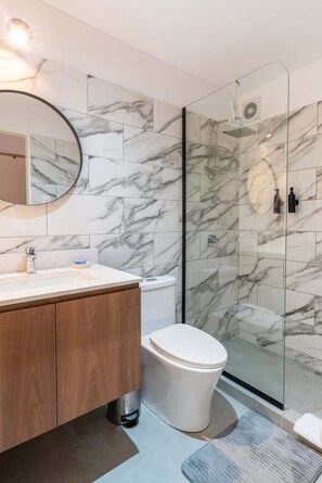 Ein modernes Badezimmer mit Marmor-Spritzschutz, einem runden Spiegel, einem hölzernen Waschtisch, einer Toilette und einer Duschkabine aus Glas.