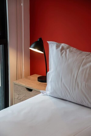 Ein Bett mit weißer Bettwäsche, einem weißen Kissen, einer schwarzen Schreibtischlampe und einem Nachttisch aus Holz.