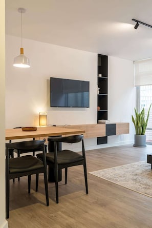 Ein modernes Wohnzimmer mit einem Holzesstisch, schwarzen Stühlen, einem an der Wand befestigten Flachbildfernseher und einem Blumentopf.