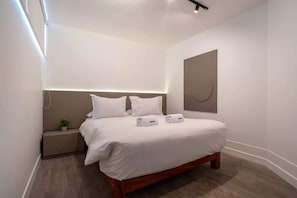 Ein modernes Hotelzimmer mit einem hölzernen Bett, weißen Leinen, einem Nachttisch mit Pflanze und einem an der Wand befestigten Kunstwerk.