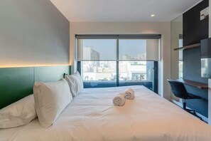 Ein Hotelzimmer mit einem großen Bett, einem Schreibtisch, einem Sessel, einem Fernseher und Blick auf die Stadt.