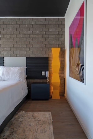 Ein Schlafzimmer mit Ziegelwand, einem Bett mit weißer Bettwäsche, einem Nachttisch, einem gelben Dekoelement und einem Bilderrahmen mit einem abstrakten Gemälde.