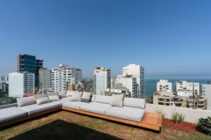 Eine Dachterrasse mit einer langen, gepolsterten Sitzgelegenheit und einem Holztisch, mit Blick auf die Stadtlandschaft und den Ozean.