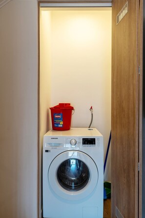 Eine weiße Samsung Waschmaschine mit einem roten Eimer darauf, platziert in einem schmalen Flur mit Holztüren.