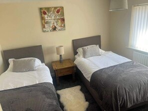 A hotel room with two single beds, a small wooden nightstand, a lamp, a painting on the wall, and a window with blinds.