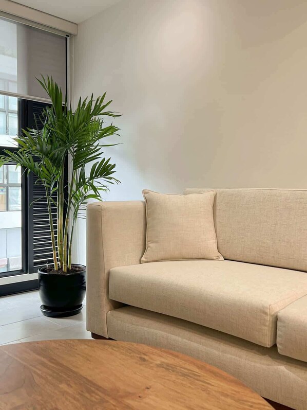 Un sofá color beige, una mesa de centro de madera y una planta en maceta en un salón moderno con un amplio ventanal.