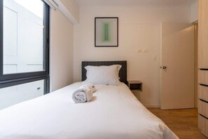 Una cama bien hecha con sábanas blancas y una toalla enrollada, una mesita de noche, un cuadro en la pared y una puerta a la derecha.