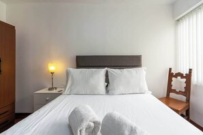 A neatly made bed with white linens, a bedside lamp, a wooden nightstand, and a chair with a decorative backrest. - Geko | Outstanding 3BR near the Malecón (Miraflores)