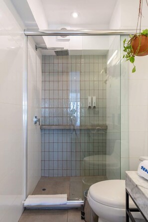 Lasiseinöillä varustettu suihku, jonka yhteydessä on laatoitettu seinä, valkoinen wc-istuin ja pieni hylly, jossa on hygieniatuotteita.