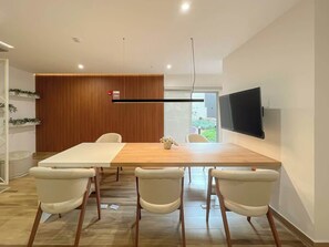 Ein zeitgemäßer Essbereich mit Holztisch, vier Stühlen, einem Wand-Flachbildfernseher und einem Holzabtrenner mit Pflanze.
