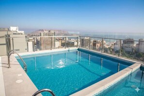 Piscina en la azotea con agua azul cristalina, rodeada por un barandal de vidrio y vistas a un paisaje urbano con edificios y una montaña lejana.