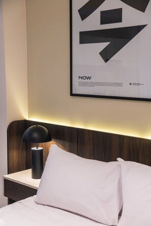 Ein modernes Hotelzimmer mit einem ordentlich bezogenen Bett, einer Nachttischlampe und einem großen, abstrakten Kunstwerk an der Wand.