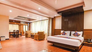 A modern hotel room with a large bed, a glass coffee table, a seating area with chairs, and a view of the outside through the window.