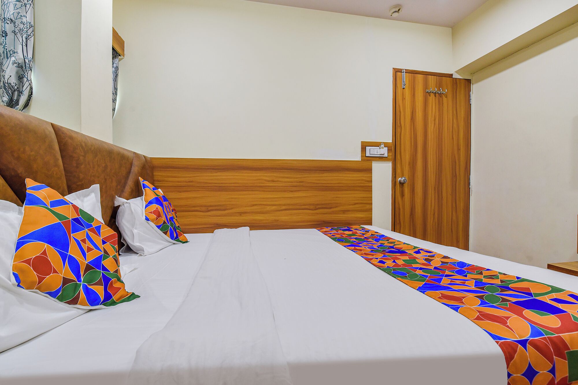 Uma cama bem arrumada com colcha estampada e almofadas, uma cabeceira de madeira e uma porta de madeira ao fundo.