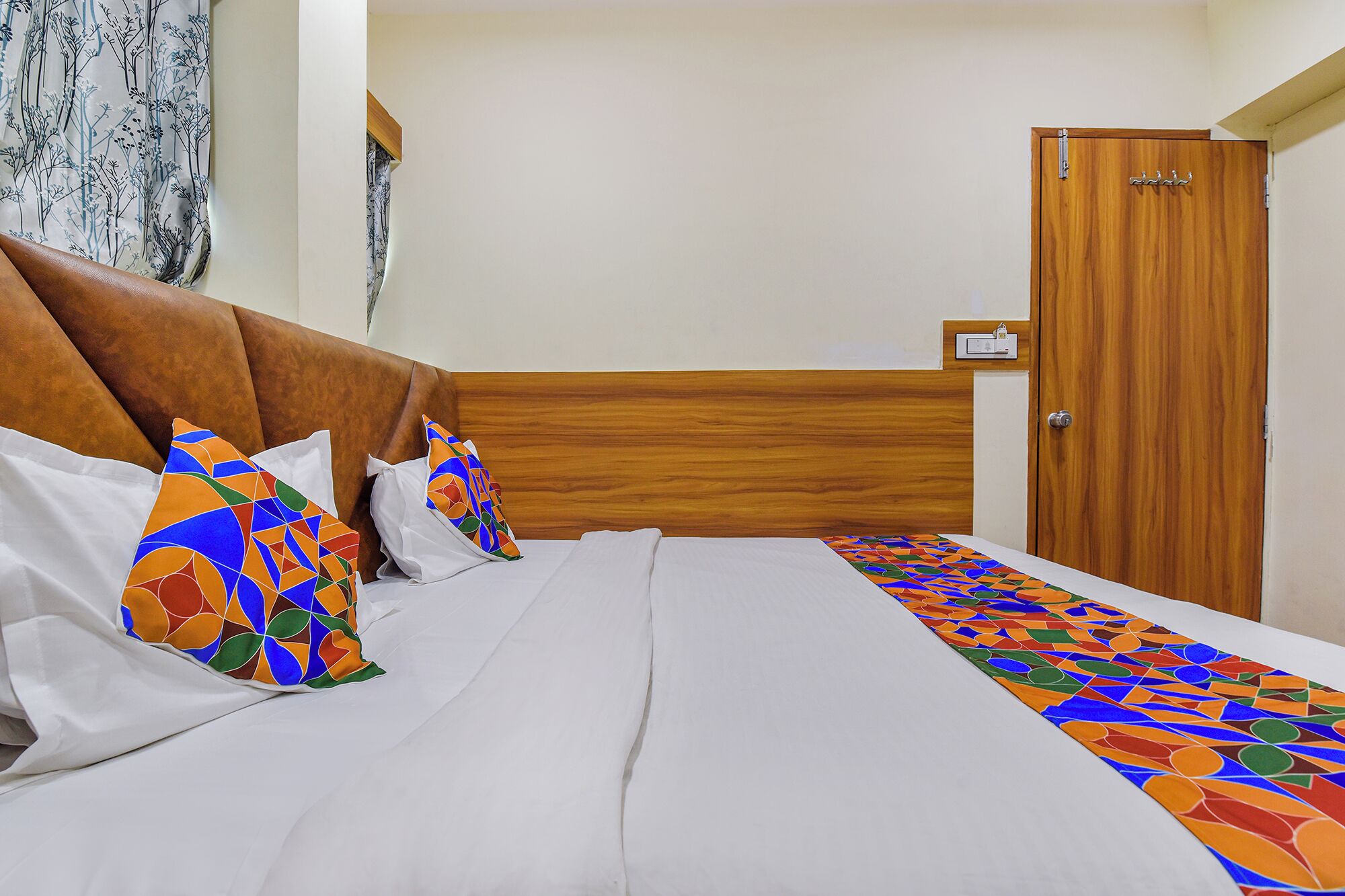 A hotel room with a wooden headboard, a bed with white linens, colorful pillows, and a patterned bed runner.