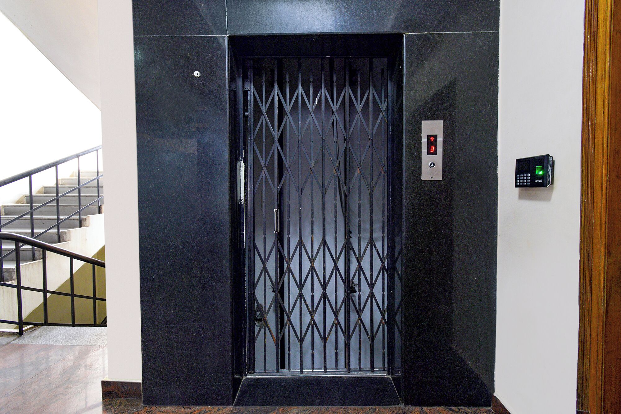 Una porta di ascensore nera con un motivo geometrico, un pannello di controllo digitale e una scala con ringhiere in metallo a sinistra.