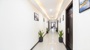 A long, well-lit hallway with potted plants on either side, leading to a door at the end.