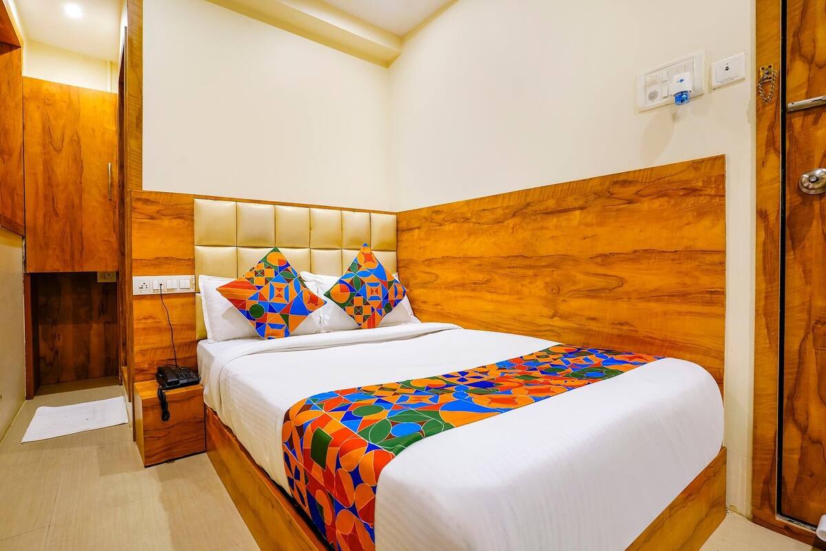 A hotel room with a wooden headboard, a bed with white linens, two colorful geometric pillows, a bedside table with a phone, and a wooden wardrobe.