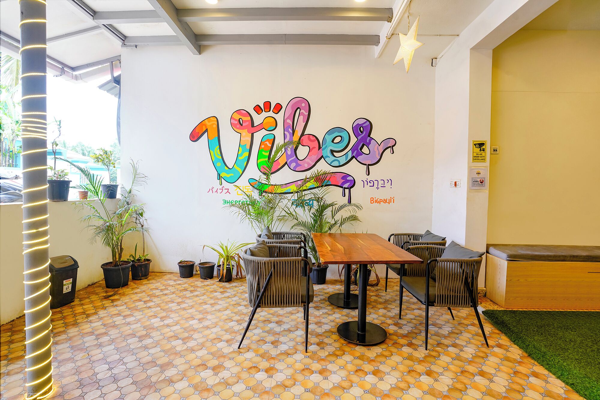A room with a tiled floor, a wooden table, and chairs. There is a wall with a colorful mural and some potted plants.