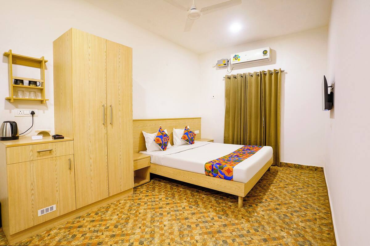 A hotel room with a bed, a wardrobe, a shelf with glasses, a kettle, and an air conditioning unit.